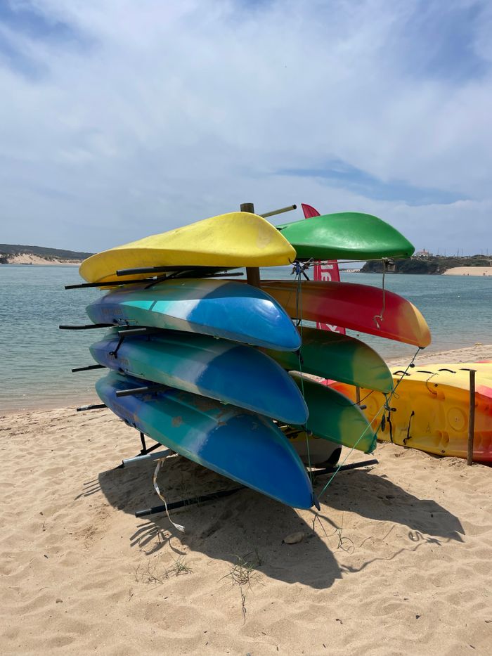 hero-img-02 Vibrant kayaks stacked on a sunny beach shore, perfect for summer adventures.